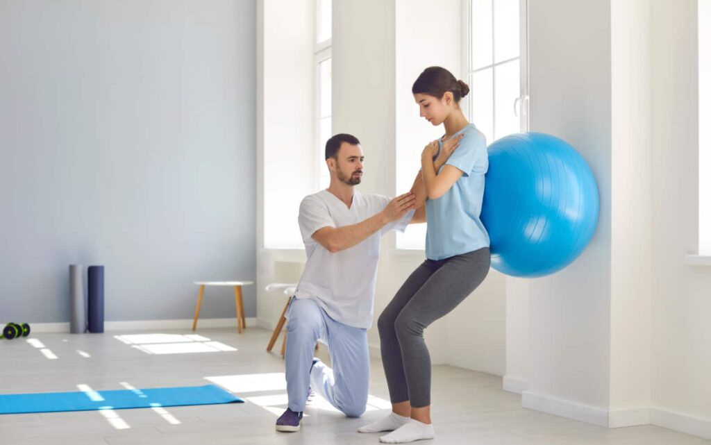 Woman performing a squat with a blue physioball with a trainer