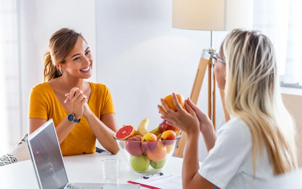 Female patient in an orange shirt meeting with a dietitian in a white shirt seated at a table.