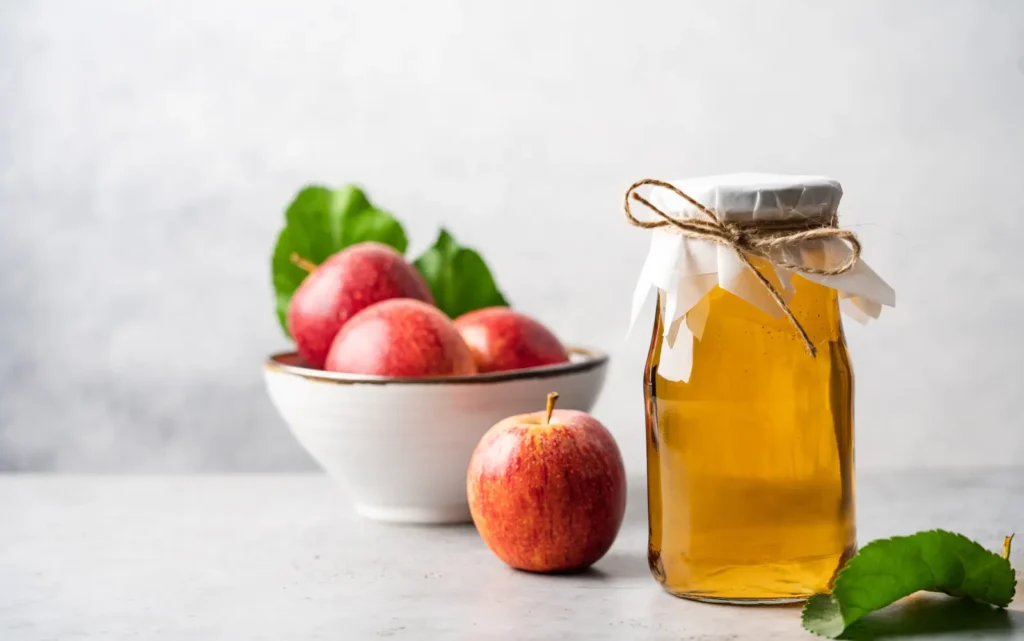 A jar of apple cider vinegar and bowl of red apples.