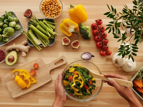 Person preparing salad off-camera with assorted fresh fruits and vegetables arranged across the table