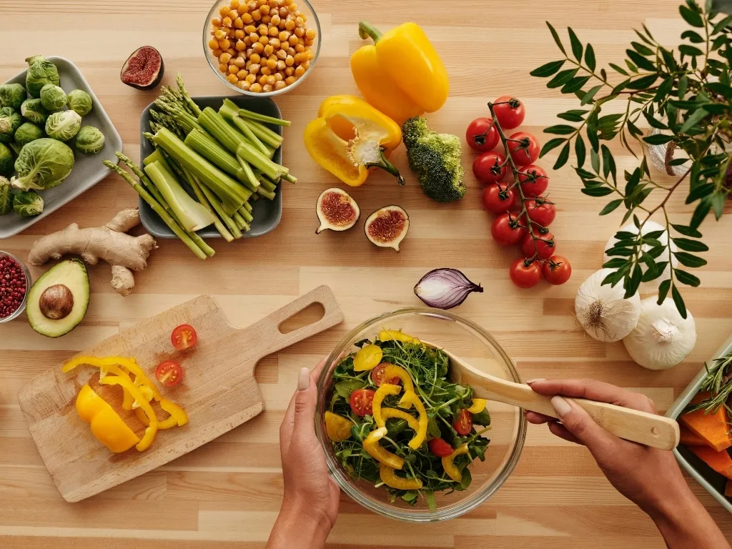 Person preparing salad off-camera with assorted fresh fruits and vegetables arranged across the table