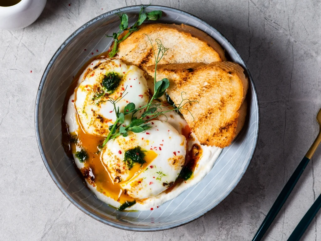 Turkish eggs served over light garlic yogurt with chili crisp and toasted sourdough, served on a blue plate alongside utensils