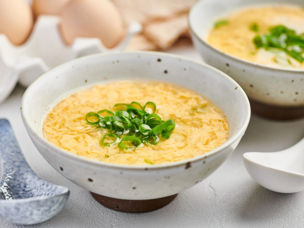 Bowls of egg drop broth with scallions, served with spoons and next to a tray of eggs