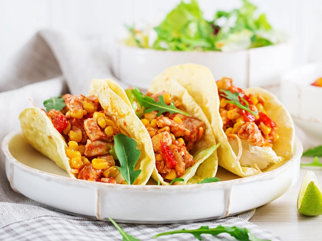 Street corn chicken tacos with lime and cabbage served on a stylish plate next to similarly designed dishware containing greens and more taco filling