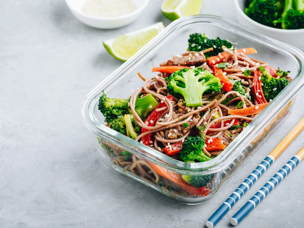 Honey sesame beef noodles with broccoli and scallions in a clear glass container next to a pair of chopsticks
