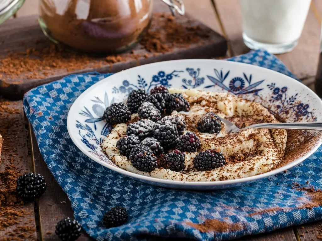 Plate of thick porridge with fresh blackberries and cocoa powder, served with a spoon on a blue tablecloth next to key ingredients