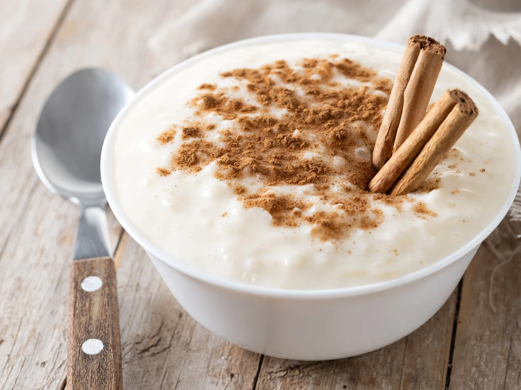 Bowl of soft rice pudding with cinnamon powder and cinnamon sticks, served on a wooden table next to a tablecloth and a spoon
