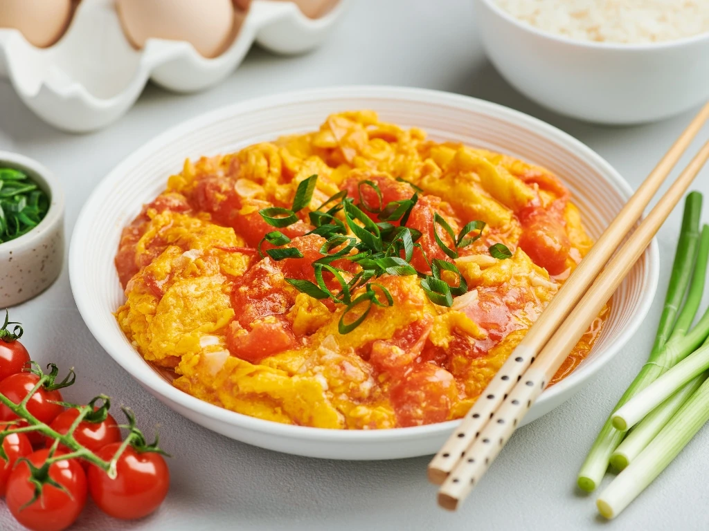 Plate of tomato and egg stir-fry garnished with green onions and served with chopsticks, next to key ingredients across the table