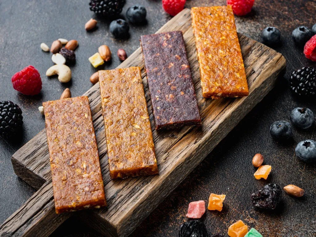 Four protein bars neatly placed on a wooden board, with fresh berries, dried fruit bits, and nuts across the table