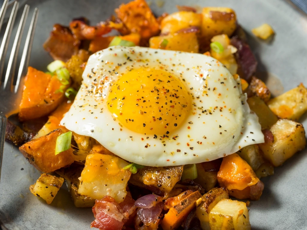 Sunny-side-up egg seasoned with pepper sits atop a savory hash of roasted potatoes, sweet potatoes, and onions