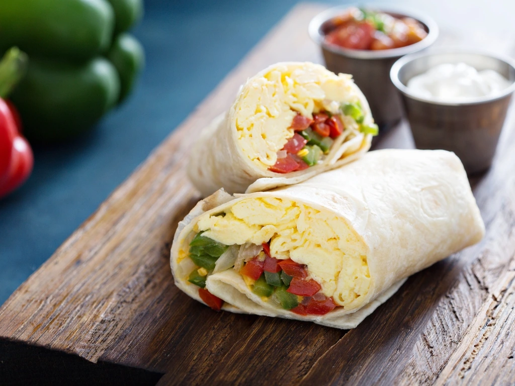 Two breakfast burritos sliced in half, filled with scrambled eggs and bell peppers, served with salsa and cream