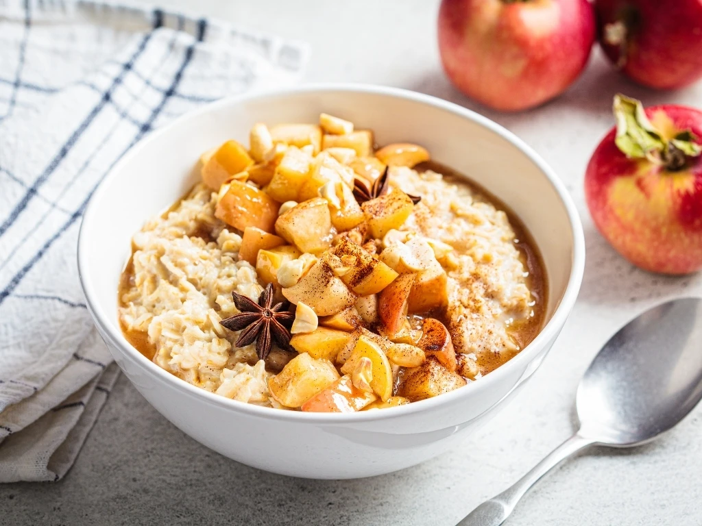 Bowl of oatmeal with sliced apple, cinnamon, and spices, next to a spoon and unsliced fruit