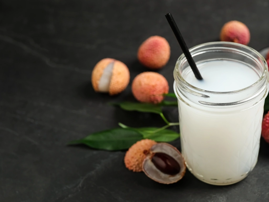 Glass jar of lychee fruit kefir with a black straw, surrounded by fresh whole and peeled lychees