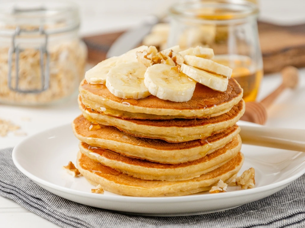 Stack of pumpkin pancakes topped with sliced bananas and nuts, with an open jar of honey, a dipper, and a jar of crushed nuts in the background
