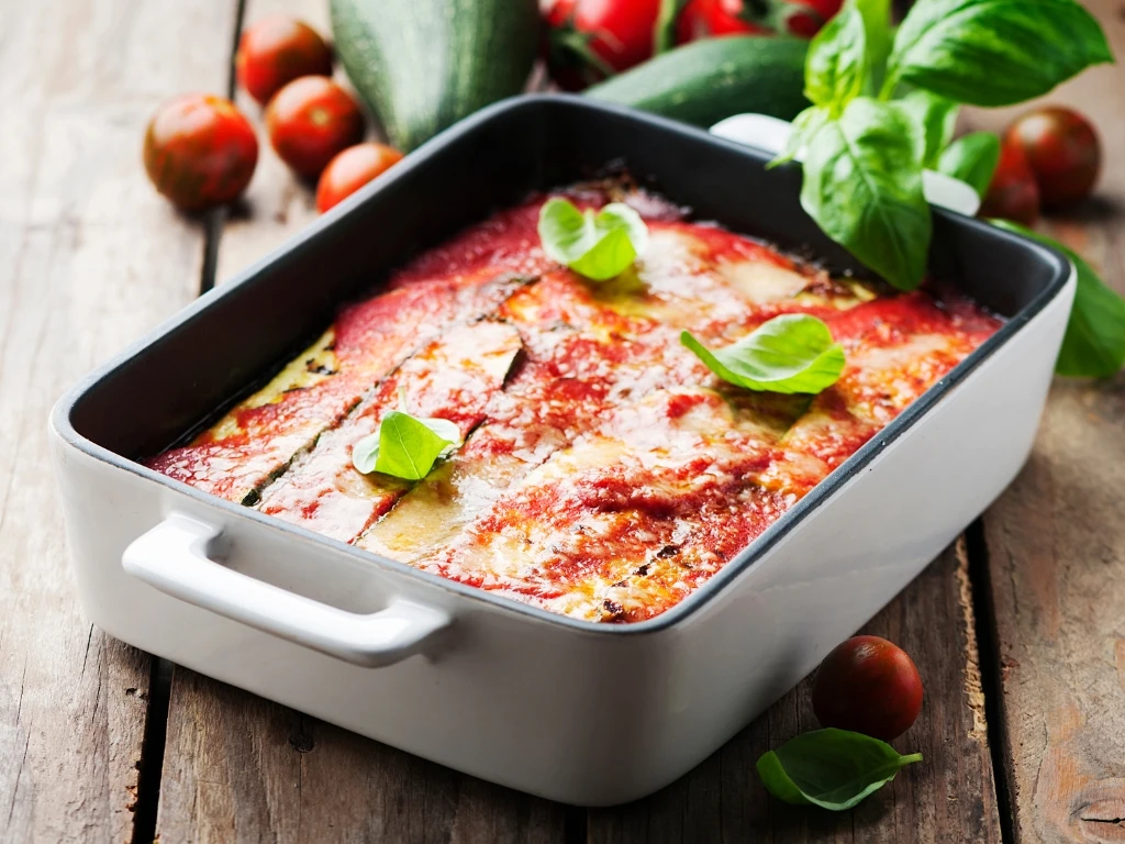 Baked zucchini lasagna in a white rectangular dish, garnished with fresh basil and served on a wooden table next to fresh ingredients