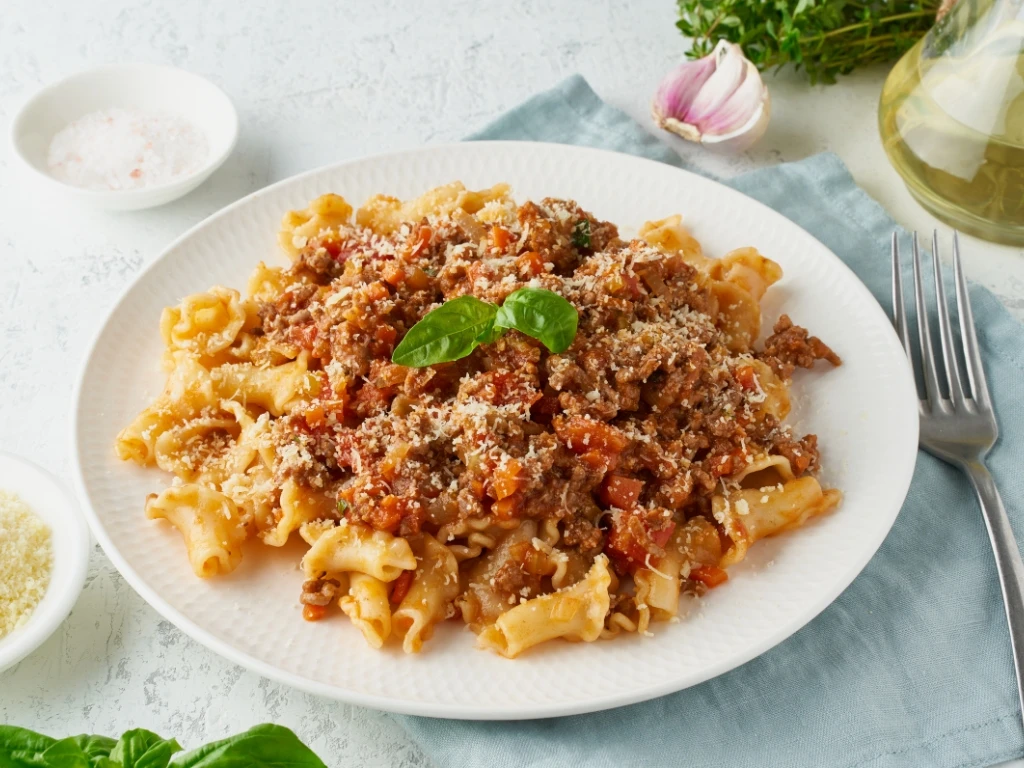 Plate of campanelle pasta topped with hearty bolognese sauce, grated parmesan cheese, and a fresh basil leaf, served with a fork next to different condiments