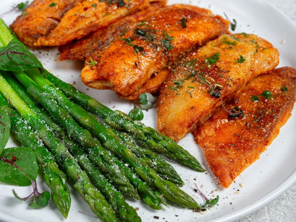 Baked fish fillet served on a white plate with a side of roasted asparagus and herbs