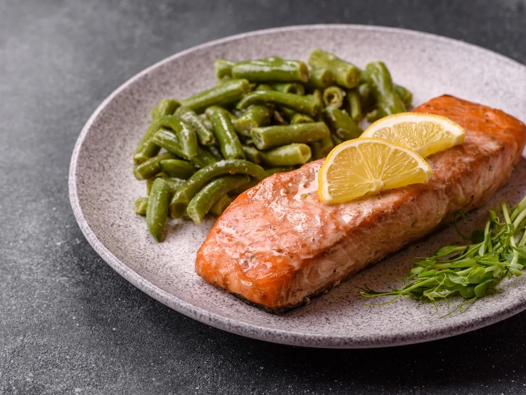 Baked salmon fillet served with lemon slices, cooked green beans, and herbs on a speckled gray plate