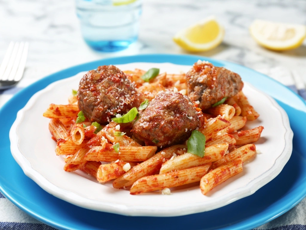 Plate of penne pasta topped with three meatballs, tomato sauce, and fresh basil leaves