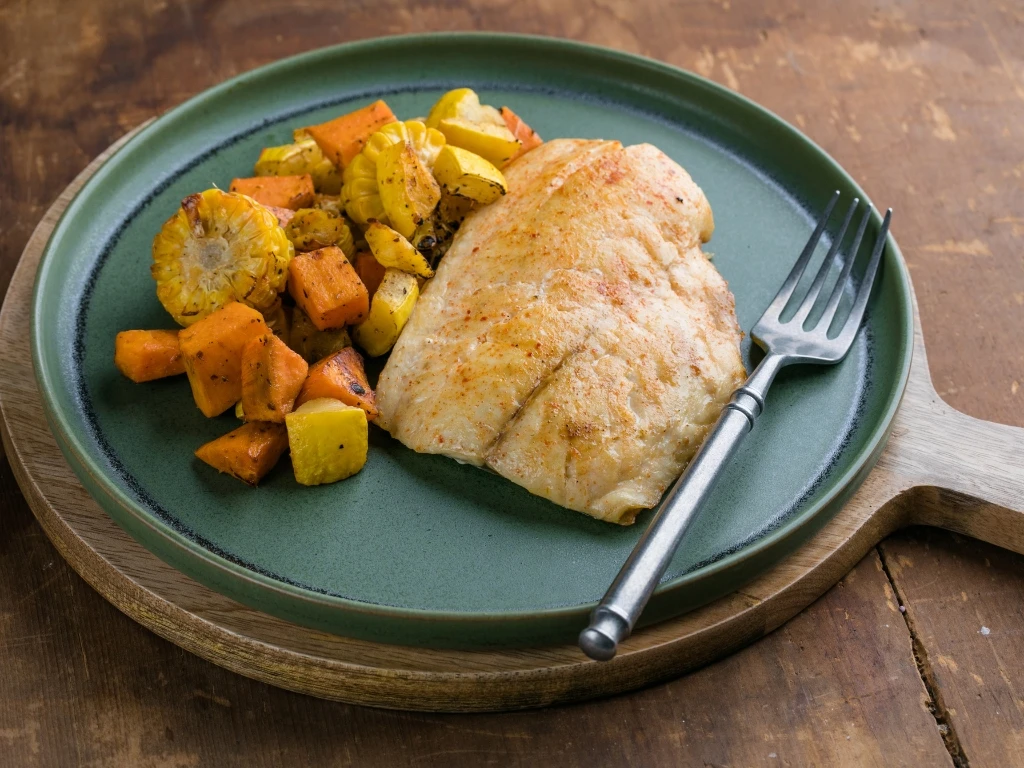 Baked cod with roasted sweet potatoes, squash, and corn, served on a blue plate with a fork
