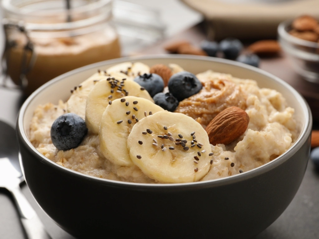 Bowl of oatmeal topped with sliced bananas, fresh blueberries, almond, almond butter, and seeds, with a spoon, a jar of nut butter, and more fruit and nuts across the table