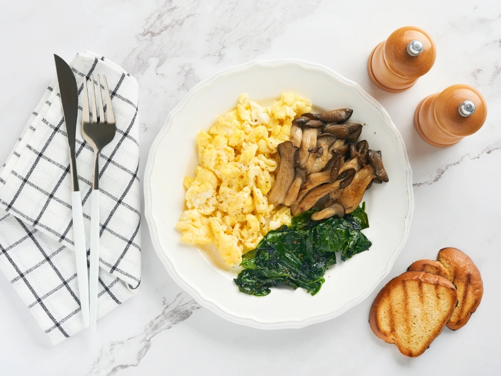 Scrambled eggs and cooked spinach and king oyster mushrooms in a white plate, served on a white marbled surface next to slices of toast, salt and pepper shakers, a tablecloth, and utensils