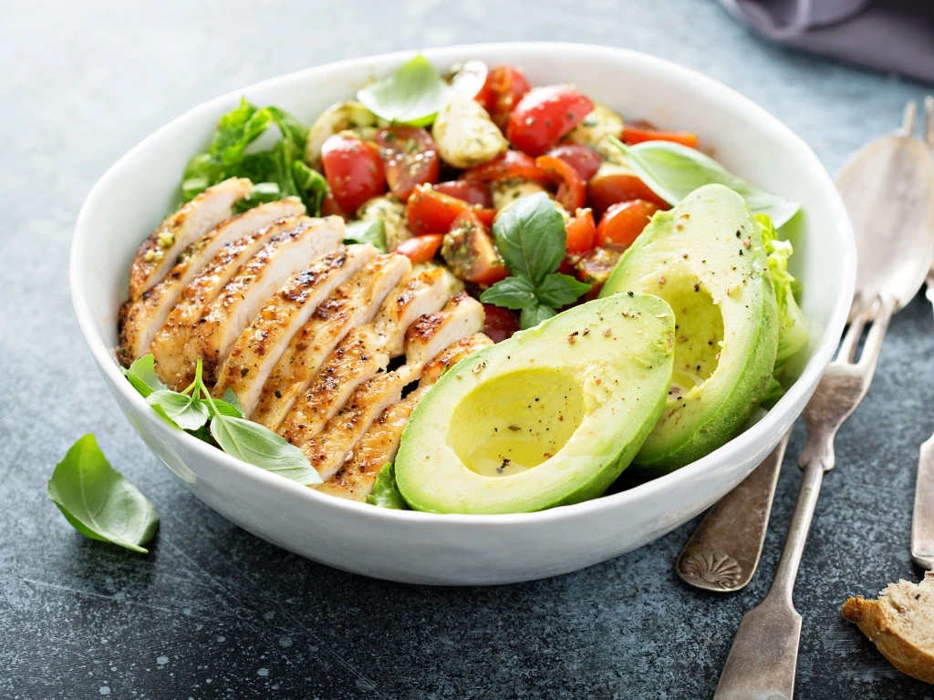 Bowl of sliced grilled chicken, red tomatoes, avocados, and leafy greens served on a dark surface with utensils