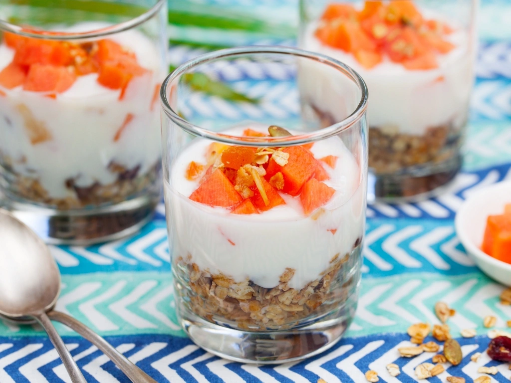 Three glasses of Greek yogurt parfait made with sliced papaya and granola, served on a playful, blue surface next to loose oats, spoons, and a small bowl of more papaya