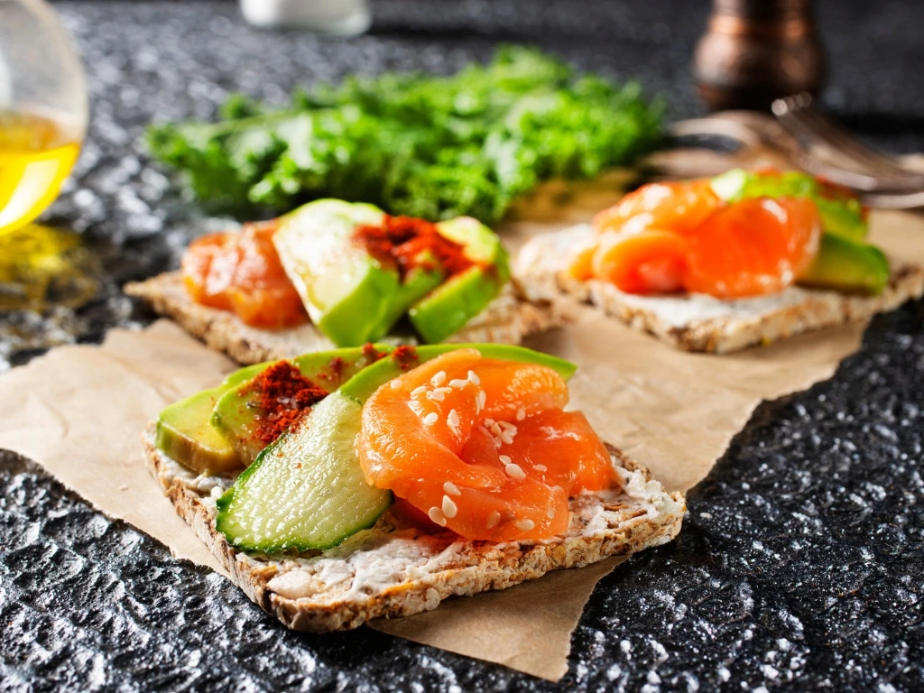 Three pieces of crispbread topped with salmon, avocado, and cucumber, with fresh herbs and a bottle of olive oil in the background