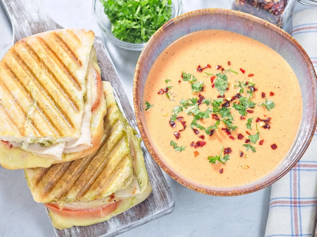 Turkey and avocado panini served on a wooden board alongside a bowl of butternut squash soup, a spoon, and some herbs