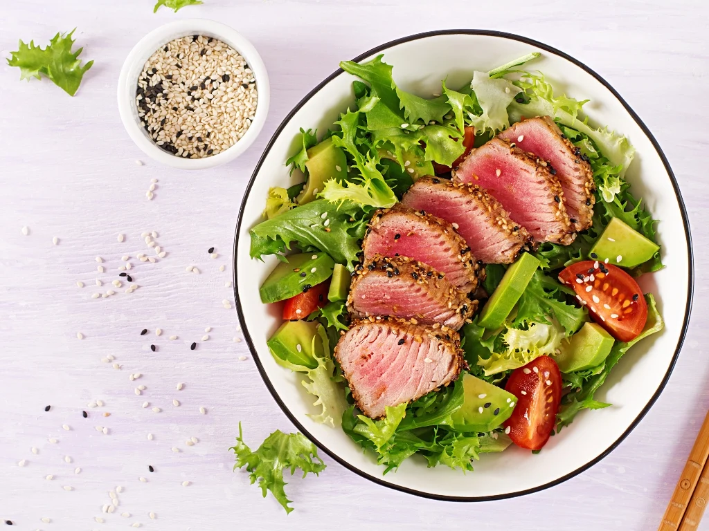 Bowl of sesame-crusted seared tuna salad with avocado, tomatoes, and mixed greens, served next to a small bowl of black and white sesame seeds