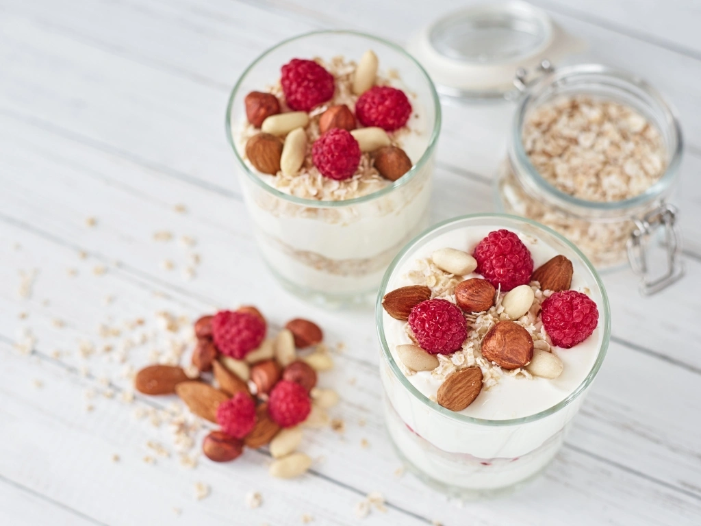Two glasses of parfait topped with nuts, oats, and berries, next to an open jar of oats and the same toppings scattered on the wooden table