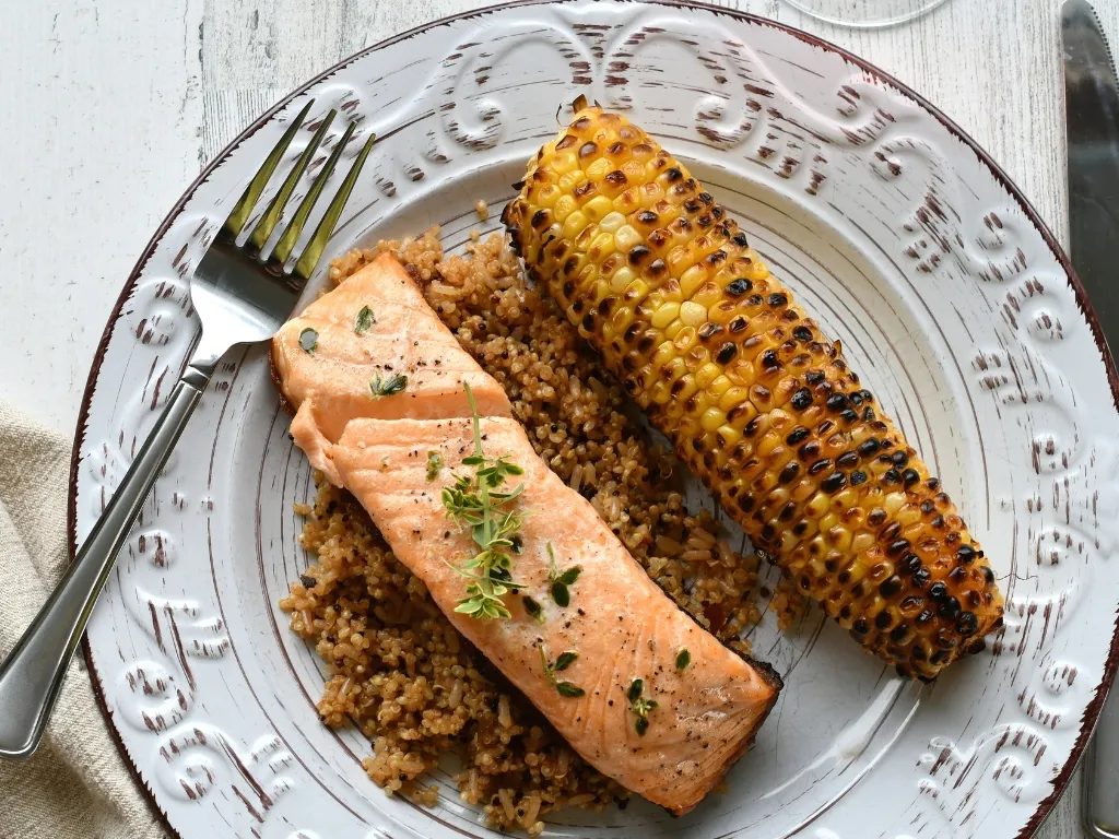 Grilled salmon with quinoa and charred corn on the cob, served on a stylish white plate alongside utensils