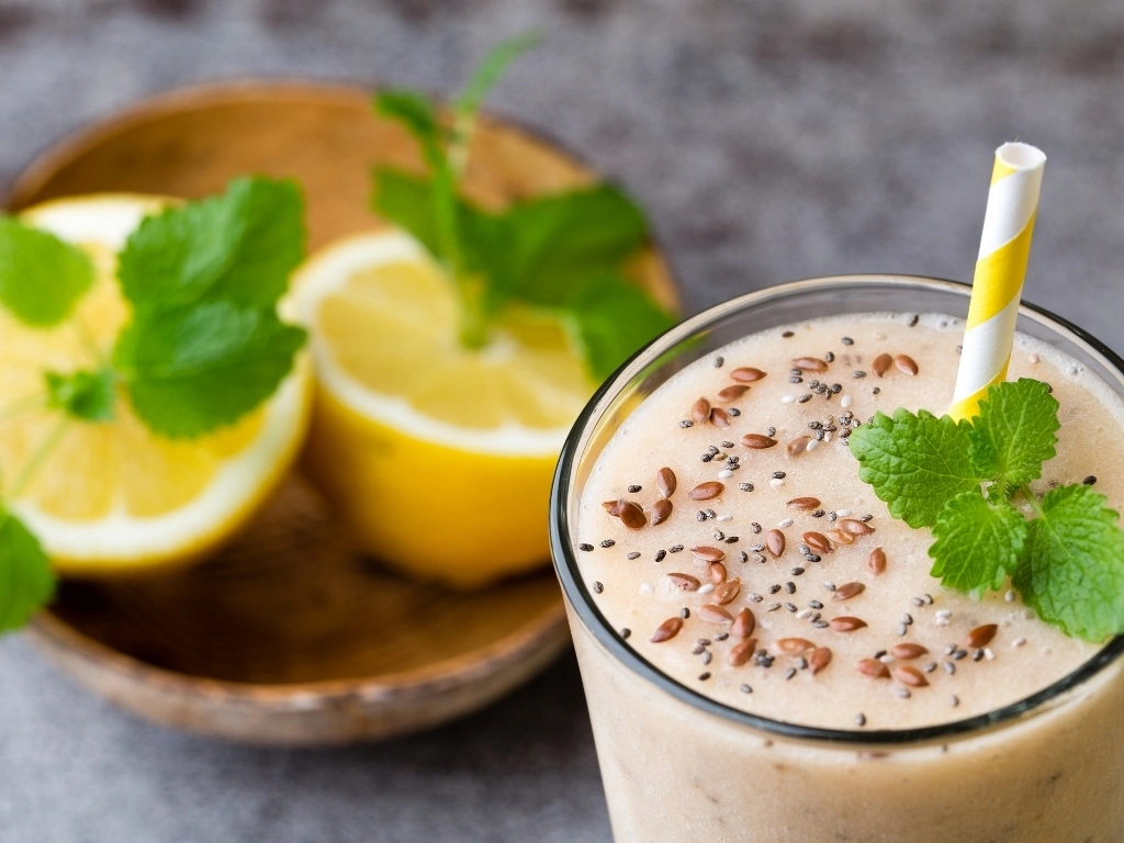 Glass of lemon smoothie garnished with mint, flaxseeds, and chia seeds, next to a wooden bowl with lemon slices