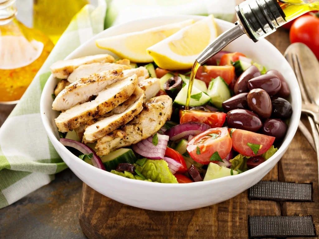 Grilled chicken salad bowl with tomatoes, cucumbers, olives, and red onion, being drizzled with olive oil