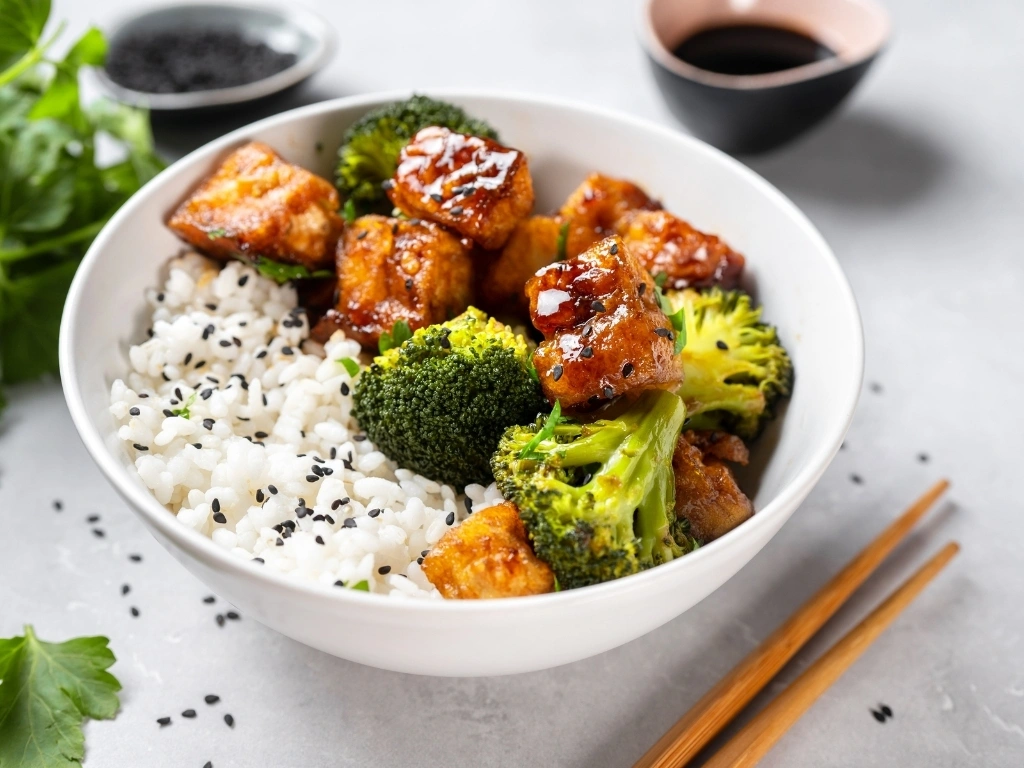 Glazed tofu and broccoli rice bowl, next to chopsticks, dips, and fresh herbs