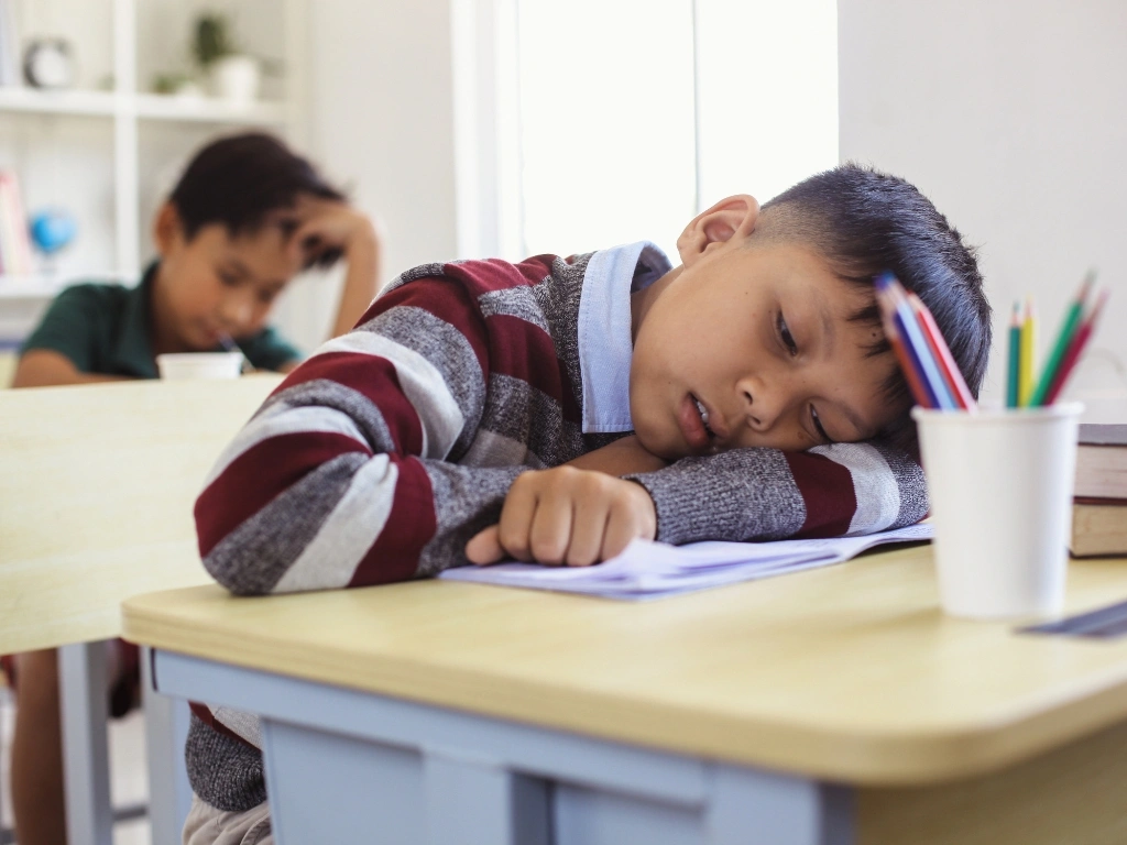 Tired student resting his head on his arms on a school desk, next to a paper cup filled with colored pencils