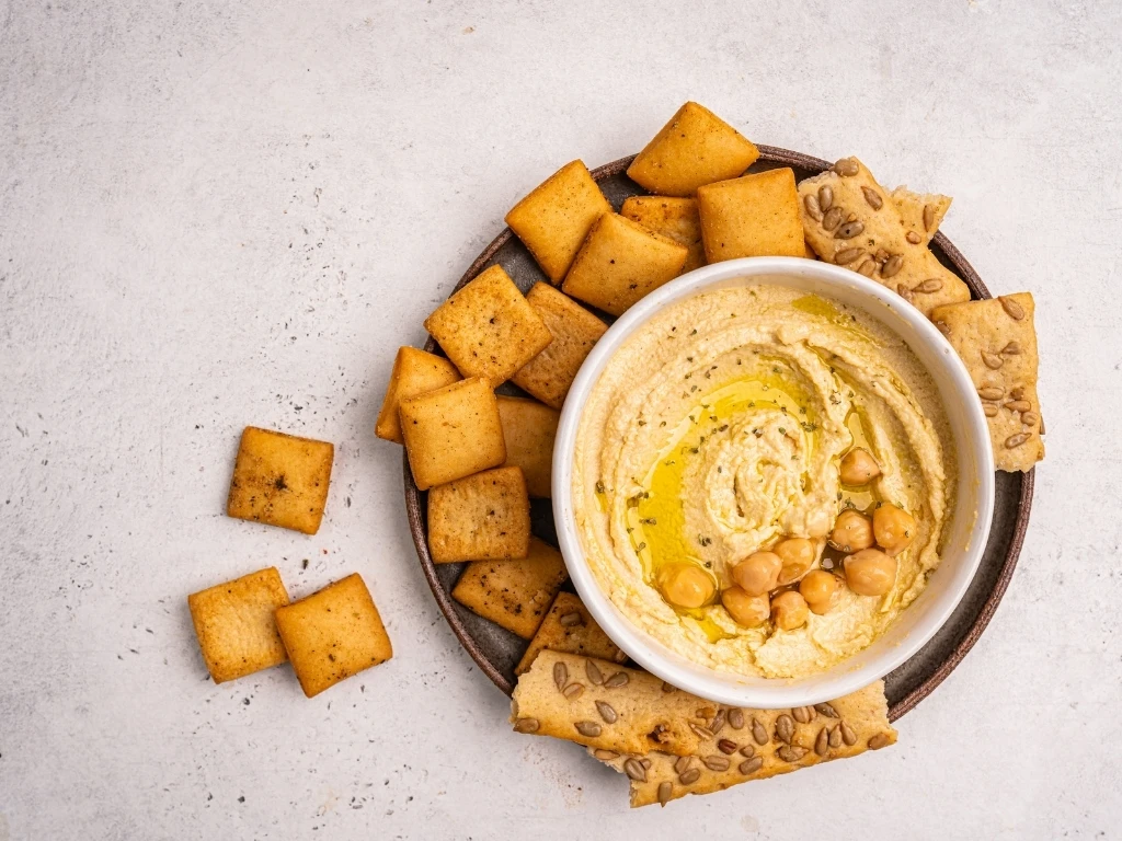 Whole-grain crackers of varied sizes and shapes on a plate, served with a bowl of hummus