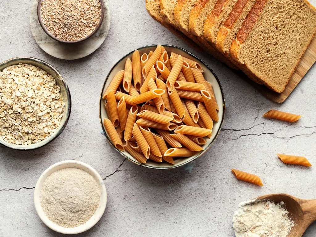 Variety of whole-grain foods, including sliced whole-wheat bread, uncooked penne pasta, rolled oats, flour, and bran, on a concrete surface