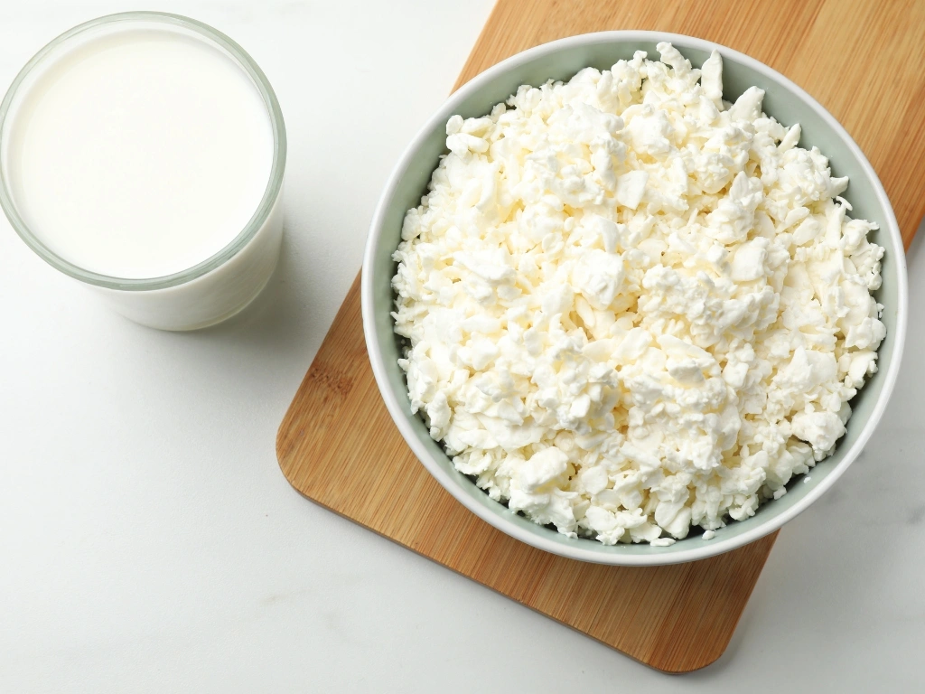Bowl of cottage cheese on a wooden board next to a glass of milk