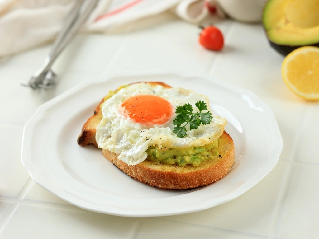 Egg and mashed avocado on toast, served on a white plate next to a fork, a slice of lemon, avocado, and tomatoes