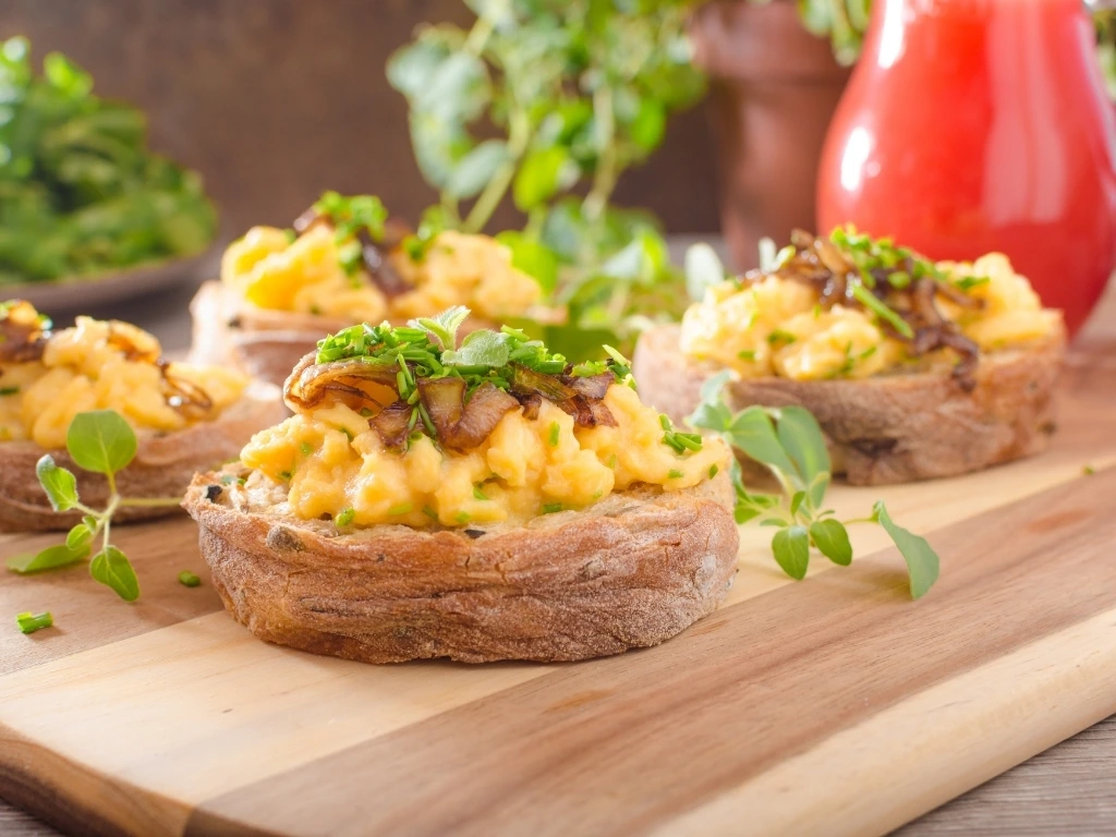 Four pieces of toast topped with scrambled eggs, chopped mushrooms, and herbs, arranged neatly on a wooden board next to a jug of juice and plate of greens