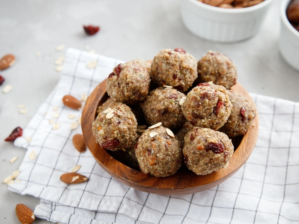 Bowl of cranberry oat balls on a tablecloth, next to ramekins of key ingredients and loose nuts and oats scattered across the table