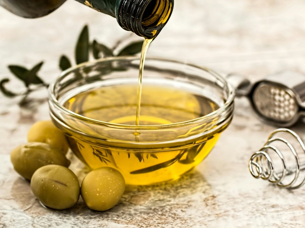 Olive oil being poured into a glass bowl surrounded by fresh olives and kitchen tools