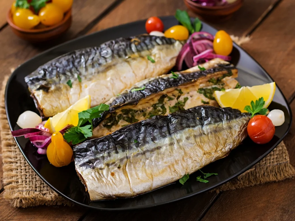 Three pieces of grilled mackerel served on a black plate and garnished with sliced lemon wedges, cherry tomatoes, parsley, and red onion