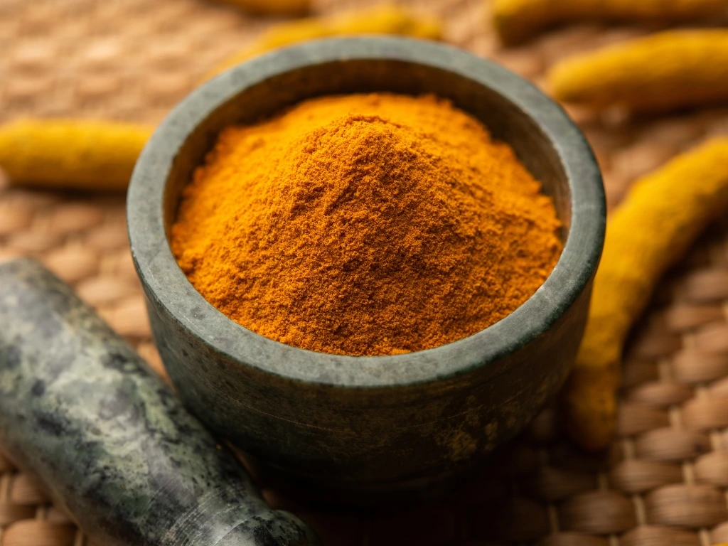 Turmeric powder in a black bowl next to a black pestle and raw turmeric roots