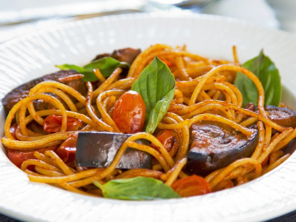 Spaghetti pasta tossed in tomato sauce with roasted eggplant, cherry tomatoes, and fresh basil leaves in a bowl