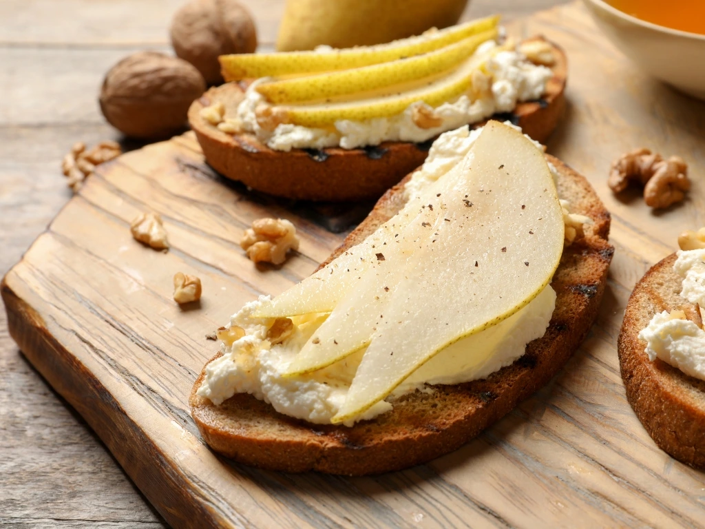 Toasted bread slices topped with creamy ricotta cheese, fresh pear slices, cinnamon, and walnuts on a wooden board