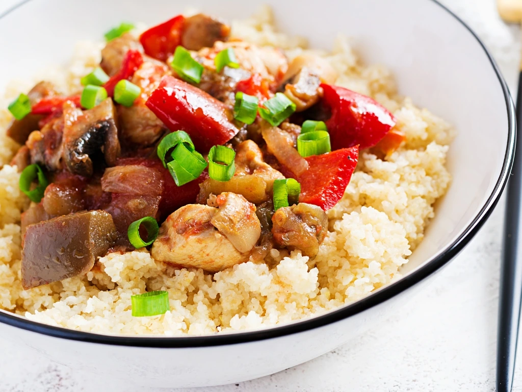 Bowl of seasoned couscous topped with stir-fried chicken and bell peppers, garnished with green onions 