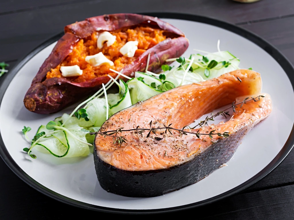 Grilled salmon steak served with a loaded sweet potato, cucumber ribbons, and fresh microgreens on a plate